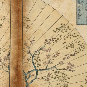 May include: A vintage Japanese folding fan with a painted design of a cherry blossom branch with pink flowers and green leaves. The fan is made of paper and has a wooden handle. The fan is open and the paper is slightly wrinkled. The fan is decorated with a pattern of small squares in a dark green color.