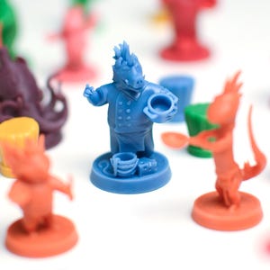 Puede incluir: Un conjunto de piezas de juego coloridas, incluyendo una figura azul con forma de chef sosteniendo una olla y una cuchara. Otras piezas son rojas, verdes, naranjas y moradas. Las piezas están sobre una superficie blanca.