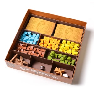 May include: A wooden box with compartments for storing game pieces. The box is brown with the words "Sör és kenyér" printed on the front. The compartments contain various colored plastic pieces, including blue, yellow, green, orange, and brown.