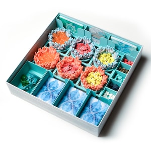 以下が含まれることがあります： ボードゲームを収納するための、ターコイズブルーの箱。箱の中には、カラフルなプラスチック製のサンゴのピース、小さなプラスチック製の魚、青と白のデザインのカードが入っています。