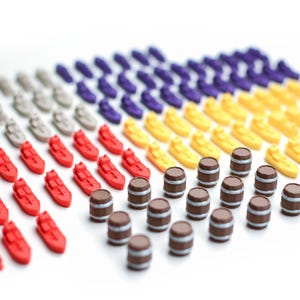 May include: A collection of miniature game pieces, including red ships, brown barrels, yellow boats, purple ships, and gray ships, arranged on a white surface. The pieces are small and detailed, suggesting a board game or tabletop activity.