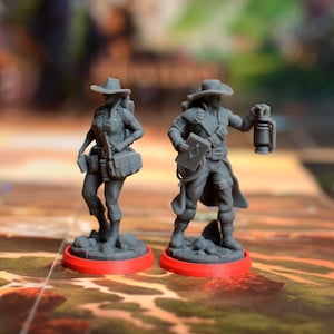 Könnte beinhalten: Zwei grau bemalte Miniaturfiguren von Abenteurern, eine weibliche und eine männliche, die auf roten Sockeln stehen. Die weibliche Figur trägt einen Hut, eine Weste und trägt einen Rucksack. Die männliche Figur trägt einen Hut, einen Mantel und trägt eine Laterne.