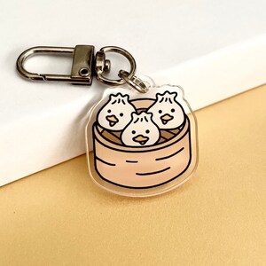 May include: A clear acrylic keychain featuring three cartoon dumplings in a bamboo steamer. The dumplings have cute faces and are arranged in a triangular shape.