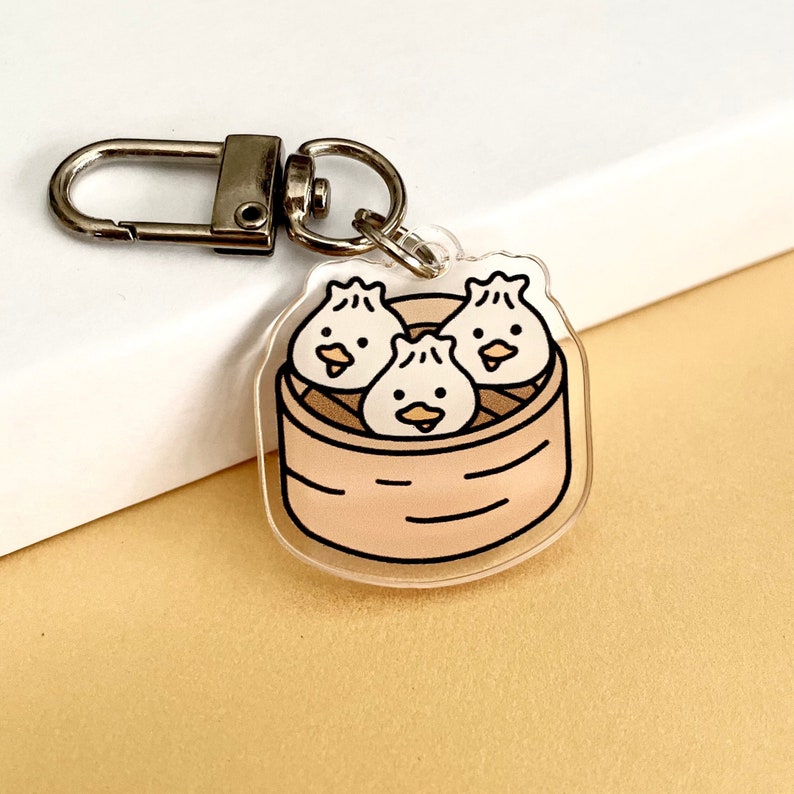 May include: A clear acrylic keychain featuring three cartoon dumplings in a bamboo steamer. The dumplings have cute faces and are arranged in a triangular shape.