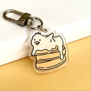 May include: A white and yellow cartoon keychain featuring a small bird with a beak sitting on top of a stack of pancakes. The keychain is made of clear acrylic and has a silver metal ring.