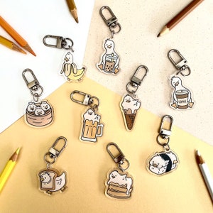 May include: Nine different keychain charms featuring a cartoon duck in various poses. The duck is holding a boba tea, a beer, a cup of coffee, a plate of dumplings, a cone of ice cream, a slice of cake, a piece of bread, and a sushi roll. The charms are made of clear acrylic and have silver key rings.