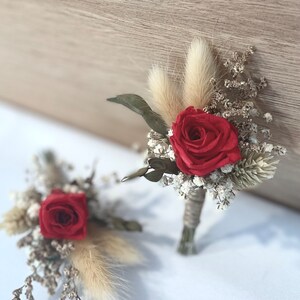 Puede incluir: Dos boutonnières de rosa roja con flores blancas secas y hierba de la pampa beige.