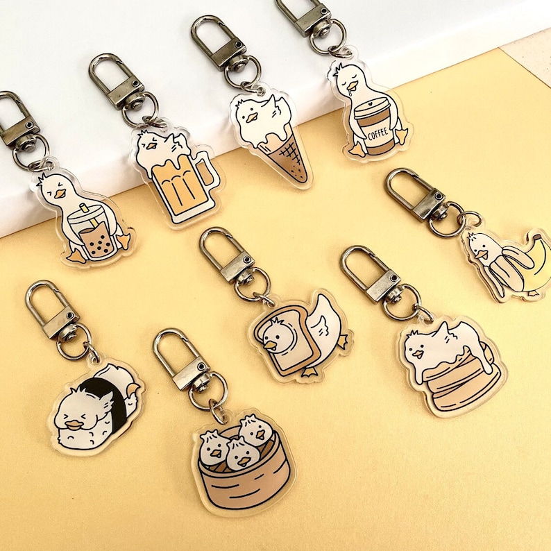 May include: Nine clear acrylic keychains featuring cartoon ducks with different food and drink items. The ducks are holding a cup of coffee, a glass of beer, a boba tea, a slice of bread, a bowl of dumplings, a banana, a cup of ice cream, a pancake, and a sushi roll.