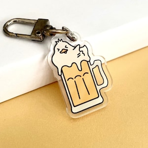 May include: A clear acrylic keychain featuring a cartoon illustration of a white chick inside a glass of beer. The chick has a yellow beak and black eyes.