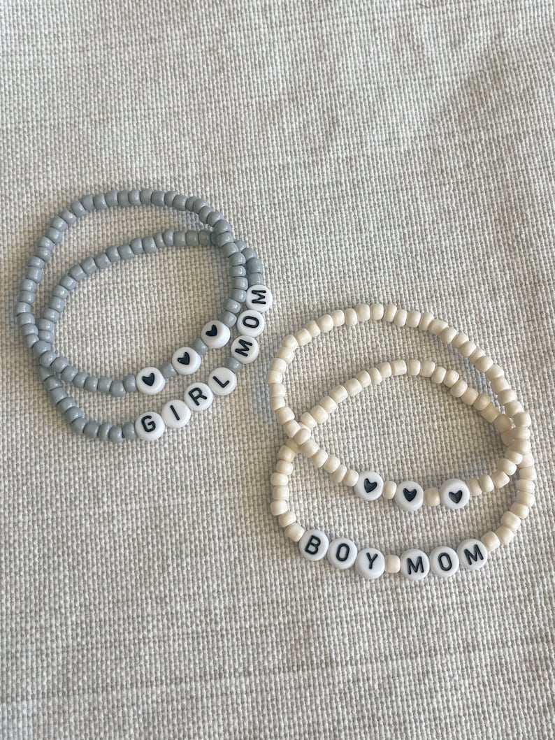 Op de afbeelding: Twee kralenarmbanden met de woorden "GIRL MOM" en "BOY MOM" erop. De armbanden zijn gemaakt van grijze en witte kralen en hebben kleine zwarte hartcharms.