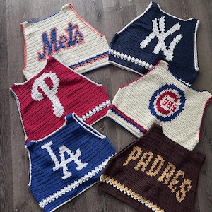 May include: Six sleeveless crochet tops featuring various baseball team logos and names, including Mets, NY, P, Cubs, LA, and Padres. Each top is crafted with distinct color schemes and textured stitching, showcasing team-specific branding in yarn.