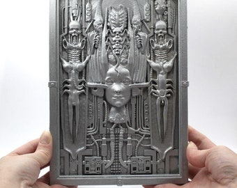 May include: A silver-colored, 3D printed wall plaque depicting a detailed, intricate design of a woman's face surrounded by biomechanical elements. The design features a central female face with a crown of intricate, biomechanical elements.
