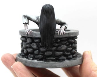 May include: A small, gray stone well with a woman figure peering out of the top. The woman is wearing a white dress and has long black hair.