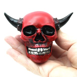 May include: Red devil skull with black horns. The skull has a toothy grin and is made of resin.