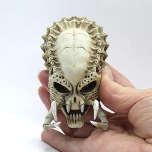 May include: A detailed, off-white skull sculpture with a textured surface and sharp teeth. The skull has a reptilian appearance with a bony ridge on the top of the head.
