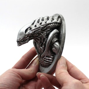 May include: A silver-coloured, detailed bust of an alien creature with a long, thin head and a textured, segmented body. The bust is mounted on a circular base.