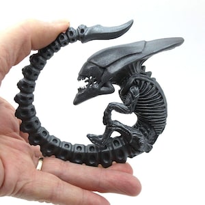 May include: A black and gray alien creature figurine with a segmented tail curled into a circle. The creature has a large head with a sharp jaw and a long, thin tail.
