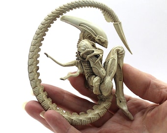 May include: A white, detailed, plastic figurine of an alien in a curled position. The alien has a long, segmented tail and a skeletal appearance.