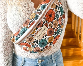 May include: A waist bag featuring a white background with a colorful floral pattern in shades of orange, yellow, and teal. The bag has a white zipper closure and a rectangular fabric label on the front with the text L'Atelier des Mirlibets.