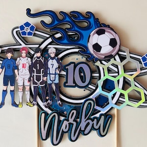May include: A layered cake topper featuring a soccer ball, blue flames, and a soccer team of four players. The number "10" is displayed, along with the name "Norbu". The design includes blue, white, and silver accents.