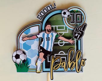 May include: A soccer-themed cake topper featuring a player in a blue and white striped jersey with the number 10. The design includes a soccer ball, the word "GOOOOL!", and the name "Pablo" in gold lettering. A #1 foam finger is also included.