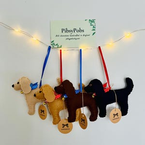 May include: Felt dog ornaments in various colors, including tan, gold, brown, and black, each with a ribbon collar and a small bell. Each dog has a handmade tag. The ornaments are suspended from blue and red ribbons.