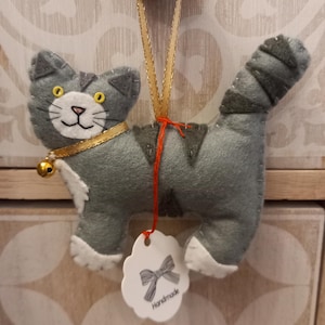 May include: A grey felt cat ornament with white accents and a gold bell. The cat has yellow eyes and a red ribbon around its neck. The ornament is hanging from a gold ribbon and has a white tag that says "Handmade".