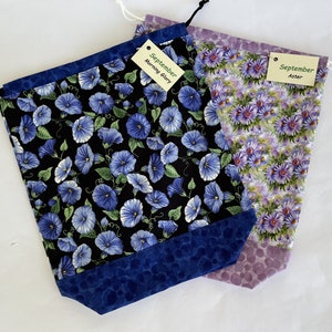 May include: Three drawstring bags with floral prints. The bags are made of fabric and have a blue, purple, and black color scheme. The bags are labeled with the month "September" and the names of the flowers: "Morning Glory", "Aster", and "Aster".
