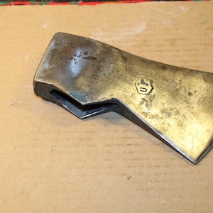 May include: A weathered metal axe head with a dark, tarnished finish and a distinct hexagonal stamp containing the letters UF. The tool features a wide, curved cutting edge and a hollow eye for a handle, showing signs of age and surface oxidation.