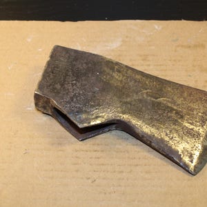 Puede incluir: Una cabeza de hacha vintage con una apariencia metálica desgastada. La cabeza del hacha tiene una hoja curva y un cuerpo rectangular. El metal tiene una mezcla de tonos oscuros y dorados, lo que sugiere edad y uso. La cabeza del hacha es una herramienta.