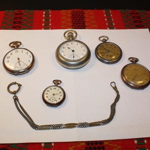 Könnte beinhalten: Eine Auswahl antiker Taschenuhren mit weißen Zifferblättern und römischen Ziffern. Die Uhren haben verschiedene Größen und Metallausführungen, einige mit aufwendigen Details. Eine Kette ist an einer Uhr befestigt.