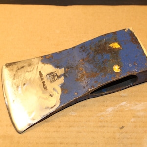 May include: A vintage axe head with a blue painted surface and a silver blade. The axe head shows signs of wear and tear, with rust and paint chipping. The words "MADE IN SWEDEN" are visible.