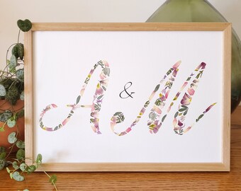 Può includere: Una stampa incorniciata con le lettere "A & M" realizzate con fiori pressati in tonalità di rosa, viola e verde. Le lettere sono in un carattere corsivo e lo sfondo è bianco.