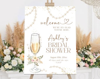 May include: A white sign with gold glitter and pearl accents announcing a bridal shower for Ashley. The sign features a champagne flute with a floral arrangement and the text "welcome, we're so glad you're here. Ashley's Bridal Shower, August 16th, 20xx, 45 days until she says 'I DO'"