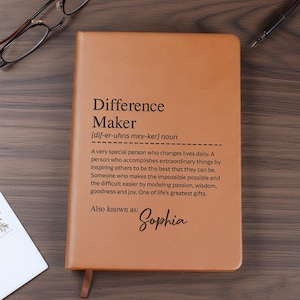 May include: A tan leather journal with the words "Difference Maker" and a definition printed on the cover. The journal also includes the name "Sophia" in cursive. The journal is lying on a wooden surface.