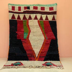 May include: A rectangular, hand-woven rug with a geometric design. The rug features a central white diamond shape, framed by red, black, and green stripes. The top border has red squares and triangles. The rug has a fringed edge.