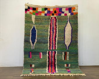 May include: Handmade green rug with a geometric design. The rug features a central vertical stripe with pink, purple, and orange accents. It has blue, white, and pink shapes, and a colorful border with squares. The rug is displayed against a white wall.