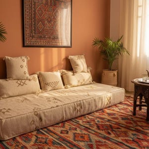May include: A cozy living room scene featuring a low, cream-colored daybed with patterned pillows. A decorative rug with a geometric design in red, orange, and blue tones covers the floor. A small wooden table holds a teapot and glasses.
