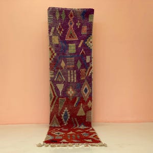 May include: A rectangular, hand-woven rug with a geometric pattern. The rug features a gradient of colors, transitioning from deep red at the bottom to purple at the top. The design includes diamond, triangle, and rectangular shapes in various colors.