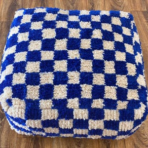 Puede incluir: Un otomán cuadrado y afelpado con un patrón de tablero de ajedrez azul y crema. La tela tiene un aspecto suave y texturizado. El otomán está colocado sobre un suelo de madera. Esta pieza decorativa se puede utilizar como reposapiés o asiento adicional.