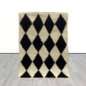 May include: A rectangular rug with a cream-colored background and a repeating pattern of black diamond shapes. The rug has a textured surface and a fringe along the bottom edge. The rug is placed on a light-colored wooden floor.