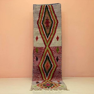 以下が含まれることがあります： 幾何学的なひし形模様の長方形の縦型ラグ。ラグは、赤、緑、黒のひし形が配置されたクリーム色の背景が特徴です。ラグはフリンジエッジで、桃色の壁に展示されています。