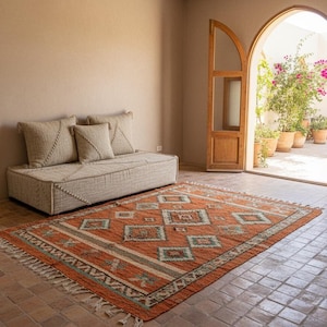 May include: An orange and green geometric patterned rug with a cream fringe sits on a tiled floor. A beige sofa with three pillows is against a beige wall. Wooden doors open to a courtyard.
