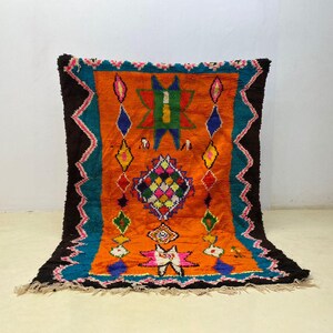 Puede incluir: Una vibrante alfombra rectangular con un campo central naranja, bordeada por turquesa y marrón oscuro. La alfombra presenta diseños geométricos en varios colores, incluyendo verde, amarillo, rosa y azul. La alfombra tiene un borde con flecos.