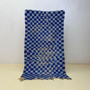 May include: A rectangular rug with a blue and grey chequered pattern. The rug has a fringe on the bottom edge and is displayed against a white wall. The rug is made of a woven material.