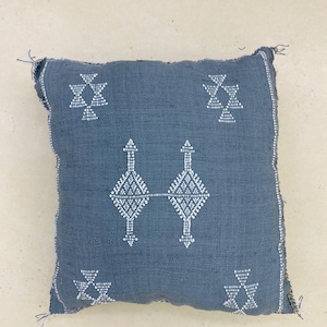 Op de afbeelding: Vierkant, blauw decoratief kussen met witte geometrische patronen. Het kussen heeft een textuurstof en een eenvoudig ontwerp. De witte patronen zijn symmetrisch en voegen visuele interesse toe.