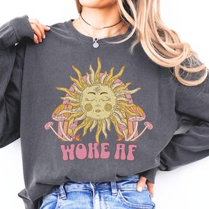 May include: A charcoal grey long-sleeved sweatshirt with a graphic of a sun with closed eyes and mushrooms. The text "woke af" is printed in pink below the graphic.