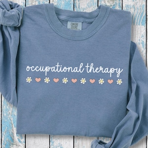 May include: A blue jean long-sleeve shirt with the words "occupational therapy" in white script. Below the text are alternating daisy and heart graphics. The shirt is made of a soft material.