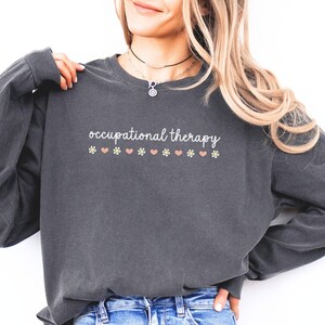 May include: A dark gray long-sleeved shirt with the text "occupational therapy" printed on the front. The text is surrounded by small pink hearts and yellow flowers.