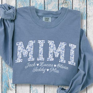 May include: A blue jean-coloured sweatshirt featuring the word "MIMI" in large, floral-patterned letters. Below, names are listed: Jack, Lucas, Olivia, Teddy, and Mia. The sweatshirt is folded on a light blue and white wooden surface.